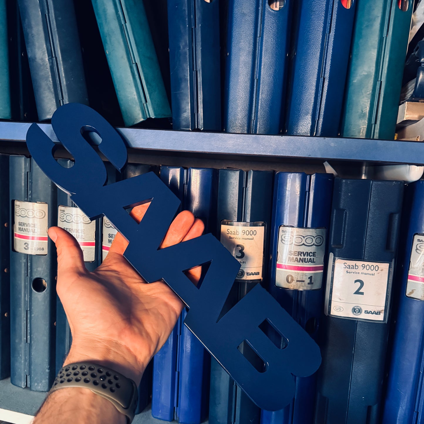 Hand holding a blue 'Saab' sign in front of blue boxes with Saab labels.
